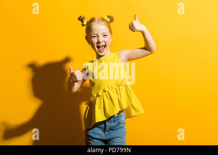 La ragazza con i capelli rossi su sfondo giallo. La ragazza ride e mostra il segno di classe. Foto Stock