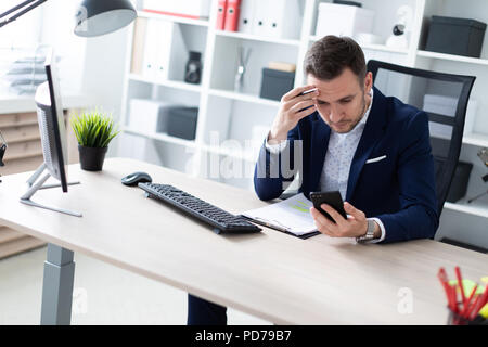 Un giovane uomo si siede in ufficio al tavolo, guarda nel telefono e tiene una matita in mano. Il giovane uomo ha documenti. Foto Stock