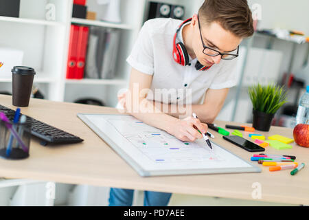 Un giovane uomo in bicchieri sorge vicino a un computer desk. Un giovane uomo estrae un indicatore su una scheda magnetica. Sul collo, il ragazzo di appendere le cuffie. Foto Stock