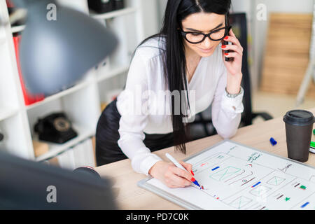 Una giovane ragazza in bicchieri sorge nei pressi di un tavolo, parla al telefono e disegna un indicatore su una scheda magnetica. Foto Stock