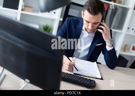 Un giovane uomo è seduto in ufficio al tavolo, parlando al telefono e tenere una matita in mano. Il giovane uomo ha documenti. Foto Stock