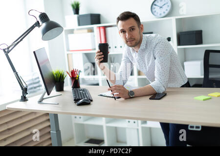 Un giovane uomo è in piedi vicino a una tabella in ufficio, tenendo in mano una matita e un bicchiere di caffè. Un giovane uomo lavora con documenti e un computer. Foto Stock