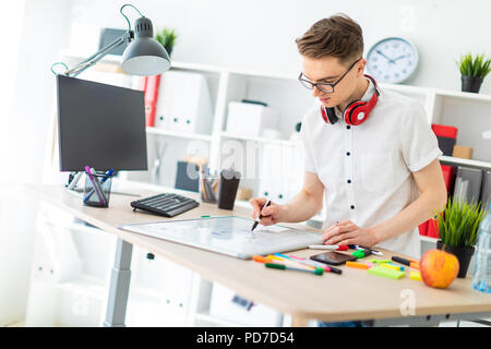 Un giovane uomo in bicchieri sorge vicino a un computer desk. Un giovane uomo estrae un indicatore su una scheda magnetica. Sul collo, il ragazzo di appendere le cuffie. Foto Stock