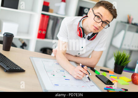 Un giovane uomo in bicchieri sorge vicino a un computer desk. Un giovane uomo estrae un indicatore su una scheda magnetica. Sul collo, il ragazzo di appendere le cuffie. Foto Stock