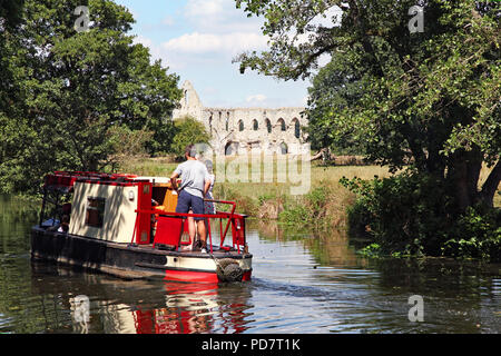 Lungo il fiume canalizzato Wey navigazione nel Surrey, Inghilterra, una chiatta sul canale trundles passato lentamente il Newark convento agostiniano con persone sul ponte godeteveli Foto Stock