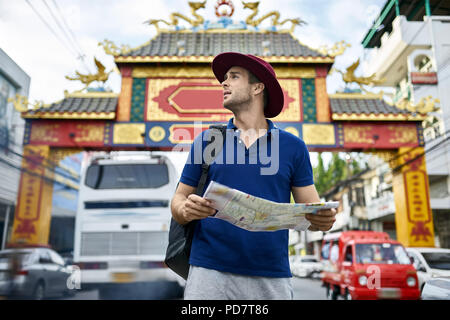 Turistico con una mappa nelle mani sorge sulla strada di città e guarda al lato. Egli indossa una polo blu, grigio pantaloncini, crimson hat e uno zaino nero. Foto Stock