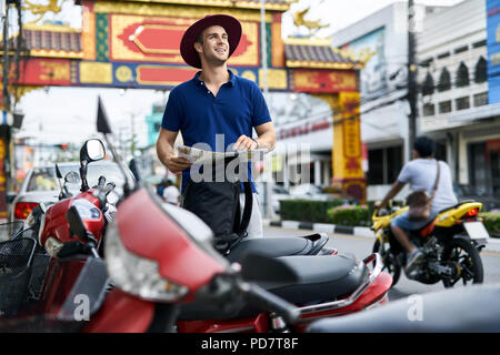 Sorridente turistico con una mappa nelle mani sorge sulla strada di città e guarda al lato. Egli indossa una polo blu, grigio pantaloncini, crimson hat e ha un blac Foto Stock