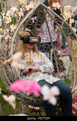 Donna che indossa le cuffie VR, seduto al posto di guida tra i fiori in realtà virtuale esperienza di orchidee - RHS Chatsworth Flower Show, Derbyshire, Inghilterra, Regno Unito. Foto Stock