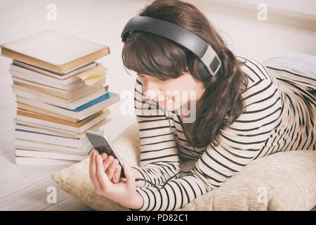 Donna l'ascolto di un audiolibro su smartphone seduto tra le pile di libri di carta sul pavimento Foto Stock