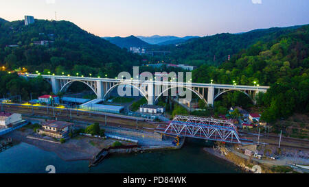 Vista Drone dell'illuminato Matsesta viadotto e ponte ferroviario sullo sfondo di montagne con una densa foresta al crepuscolo, Sochi, Russia Foto Stock