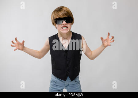 Ritratto di adorabili funnylittle boy in piedi in studio con occhiali da sole che mostra agression rabbia che vogliono graffio su sfondo grigio Foto Stock