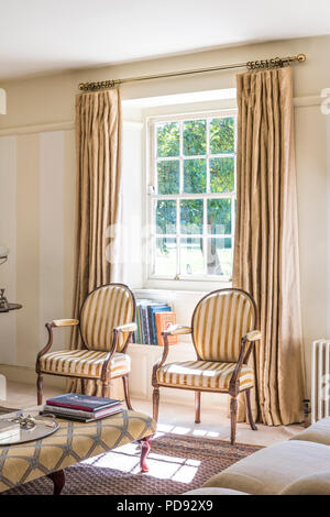Coppia di strisce di stile antico sedie nella finestra della stanza di seduta con stile Regency ottomano e biancheria lunghezza pianale tende Foto Stock