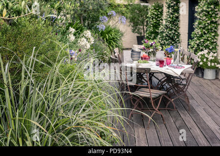 Vecchio bistrò francese sedie intorno a lungo tavolo in legno su giardino terrazza sul tetto con impalcati. Agapanthus e gelsomino fiorisce in background Foto Stock