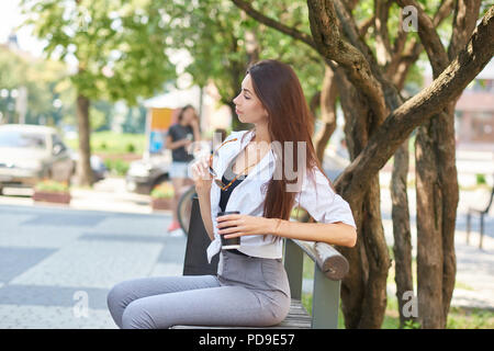 Elegante ragazza seduta sul banco in downtown, mantenendo il quartiere alla moda di occhiali. Modello di colore grigio che indossa pantaloni classici, black tshirt, camicia bianca. Alberi verdi Foto Stock