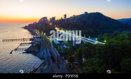 Drone vista del mare con la ferrovia, illuminata Matsesta viadotto e le montagne con una densa foresta al crepuscolo, Sochi, Russia Foto Stock