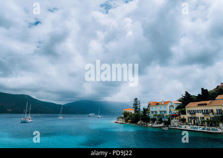 Cloudscape oltre Fiskardo village edifici con mattoni di colore arancione di tetti. Yacht barche che cercano rifugio nella baia. L'isola di Cefalonia in Grecia Foto Stock