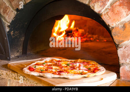 Rustico pizza fatta in casa il prosciutto cotto in forno a legna per la stufa in mattoni con fuoco in background Foto Stock