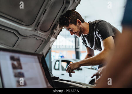 Sorridente giovane uomo che ripara la macchina in garage con un collega utilizzando laptop nella parte anteriore. Lavoro meccanico sotto il cofano di un veicolo. Foto Stock