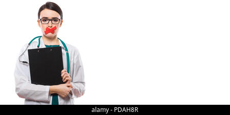 Giovane donna medico con stetoscopio negli appunti di contenimento nelle sue mani con labbra con nastro in divisa bianca su sfondo bianco Foto Stock