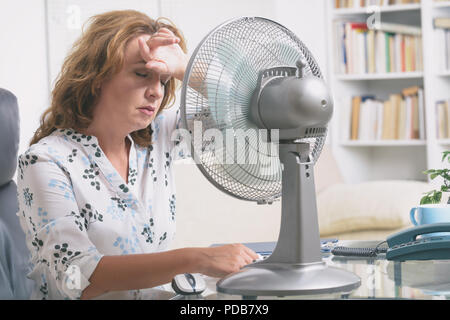 La donna soffre di calore mentre si lavora in ufficio e si tenta di raffreddarsi dalla ventola Foto Stock