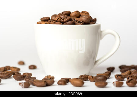 Porcellana Bianca tazzina su sfondo bianco riempito di caffè torrefatto in grani che hanno versato sulla superficie Foto Stock
