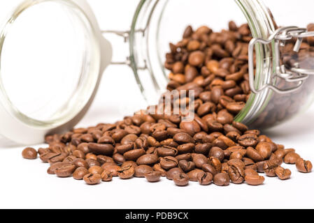 Vetro storage alimentare jar giacente sul lato con i chicchi di caffè riversandosi su superficie bianca Foto Stock