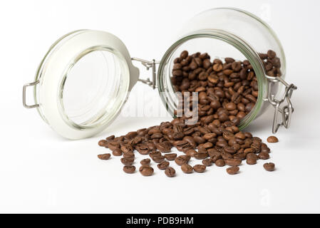 Vetro storage alimentare jar giacente sul lato con i chicchi di caffè riversandosi su superficie bianca Foto Stock