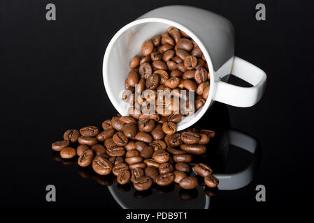 Chicchi di caffè tostati fuoriuscita dal bianco tazzina giacente sul lato nero riflettente superficie di vetro Foto Stock