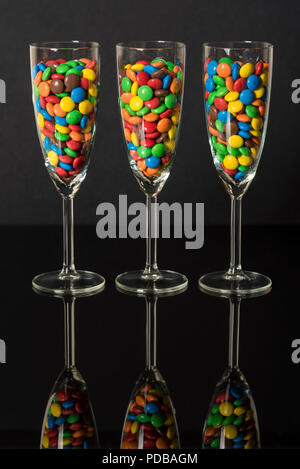 Rivestite di zucchero caramelle di vari colori in tre flauti champagne su uno sfondo nero con la riflessione Foto Stock