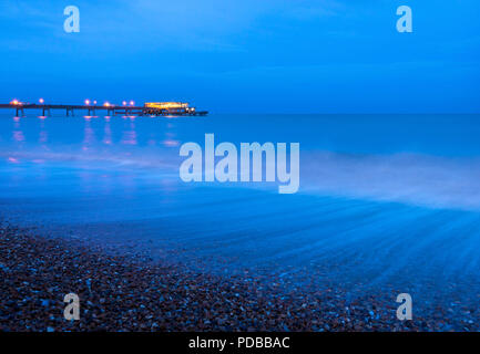 Onde che si infrangono lungo il lungomare di trattativa al tramonto con il Molo di trattativa illuminato in background. Foto Stock