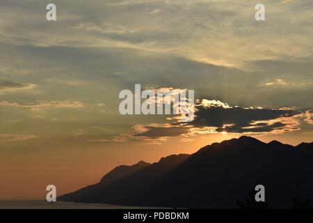 Arancio brillante tramonto scontornamento le montagne della costa della Dalmazia Croazia con la luce del sole raggiante attraverso le nuvole Foto Stock