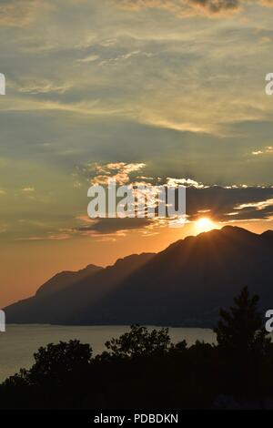 Arancio brillante tramonto scontornamento le montagne della costa della Dalmazia Croazia con la luce del sole raggiante attraverso le nuvole Foto Stock