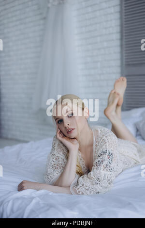Giovane e bella sposa in negligee giacente sul letto e guardando la fotocamera Foto Stock