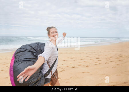 Ritratto sorridente e spensierato giovane femmina di parapendio con zaino paracadute su Ocean Beach Foto Stock