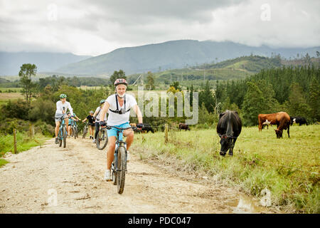 L'uomo mountain bike con gli amici su rurale strada sterrata lungo il pascolo di vacca Foto Stock