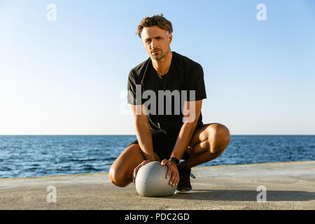 Bel uomo adulto seduto con montare la sfera sulla riva del mare e guardando la fotocamera Foto Stock