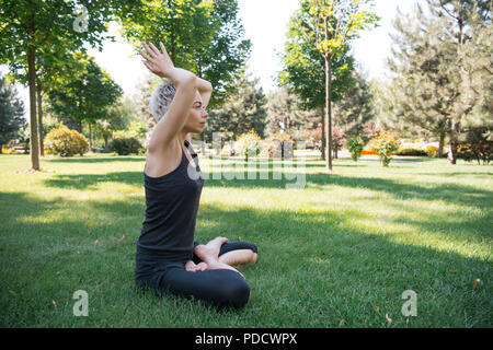 Vista laterale della donna a praticare yoga in lotus posano con le mani alzate rendendo namaste gesto su erba nel parco Foto Stock