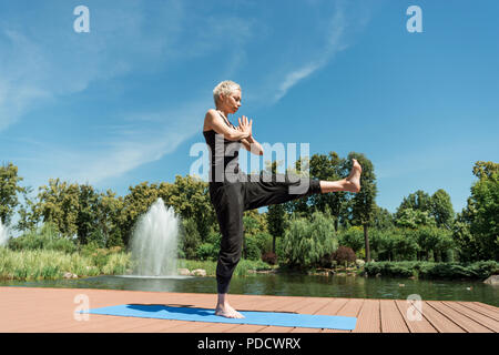 Vista laterale della donna a praticare yoga sul materassino yoga e rendendo namaste gesto vicino al fiume in posizione di parcheggio Foto Stock