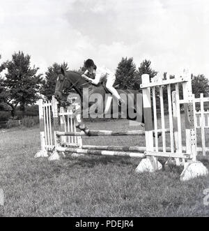 Anni sessanta, una ragazza adolescente in sella ad un cavallo salta un obstable o recinzione in un campo, Inghilterra, Regno Unito. Indossare un casco e mostra buona tecnica, sul cavallo, ella è mettere in pratica la sua abilità di showjumping. Foto Stock