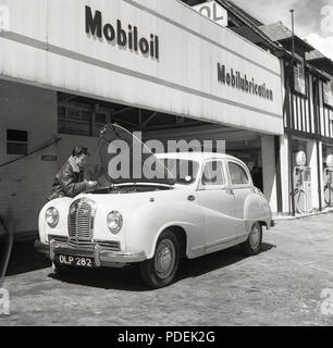 Degli anni Cinquanta, storico, un'auto Austin con il suo cofano motore fino parcheggiata fuori un Mobil stazione di servizio, un giovane uomo in una giacca di volo il controllo dell'auto olio motore, Inghilterra, Regno Unito. Foto Stock