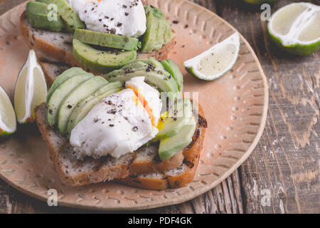 Uovo affogato e fette di avocado su pane integrale tostato servito con calce sulla piastra in ceramica su tavola in legno rustico, panini vegetariani Foto Stock