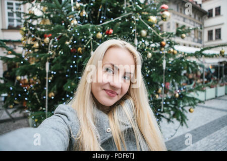 Una giovane e bella donna bionda o ragazza facendo selfie accanto a un albero di Natale durante le vacanze di Natale in Piazza della Città Vecchia di Praga, Repubblica Ceca. Foto Stock