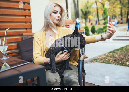 Bella e felice donna bionda godendo in cafe bar e prendendo selfie foto con la sua adorabile bulldog francese. Foto Stock