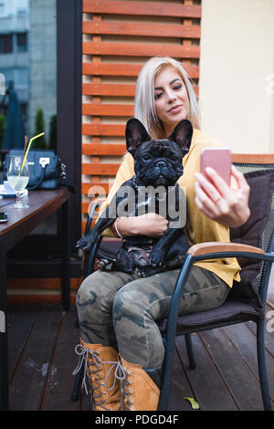 Bella e felice donna bionda godendo in cafe bar e prendendo selfie foto con la sua adorabile bulldog francese. Foto Stock