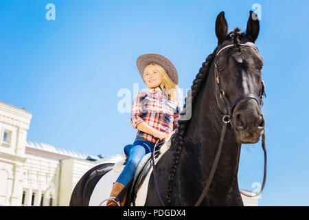 Ragazza sorridente indossano jeans e stivali da cavallo seduto sul cavallo scuro Foto Stock