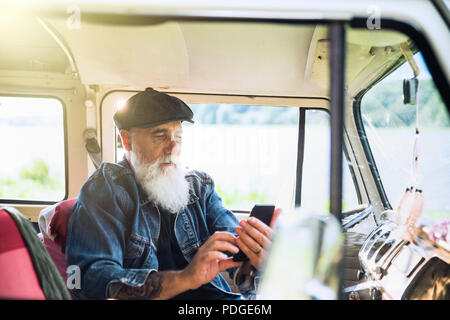 Un senior hipster seduta nel suo camper, utilizzando un telefono cellulare. Foto Stock