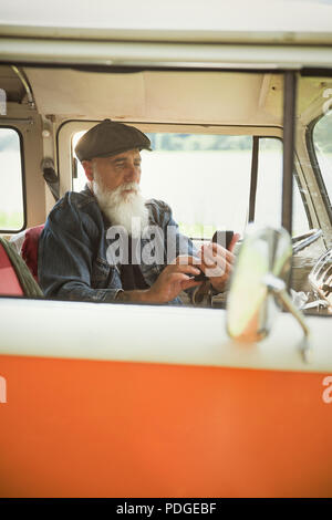 Un senior hipster seduto nel suo furgone vintage, utilizzando un telefono cellulare. Foto Stock