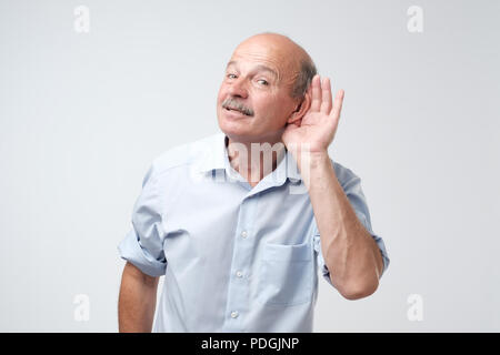 Ritratto di senior uomo informale che overhears conversazione su sfondo bianco. Parlare ad alta voce si prega di concetto. Che cosa hai detto Foto Stock