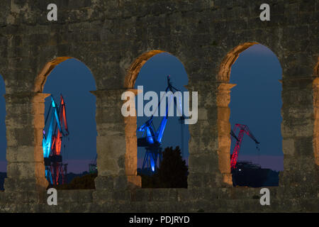 Rovigno Croazia - Luglio 31, 2018: vista notturna attraverso le arcate dell'anfiteatro di Pula al illuminato gru del porto della città croata. Foto Stock