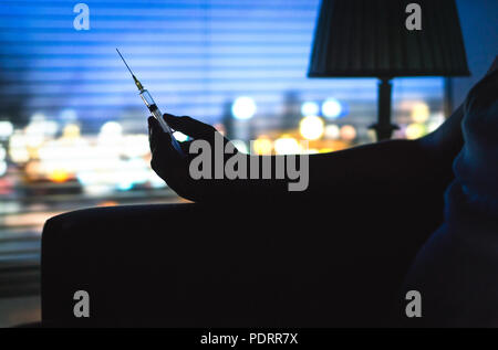 Tossicodipendente con siringa di eroina silhouette nel buio. Mano azienda ago a tarda notte. Luci della città lo sfondo da home appartamento finestra. Foto Stock
